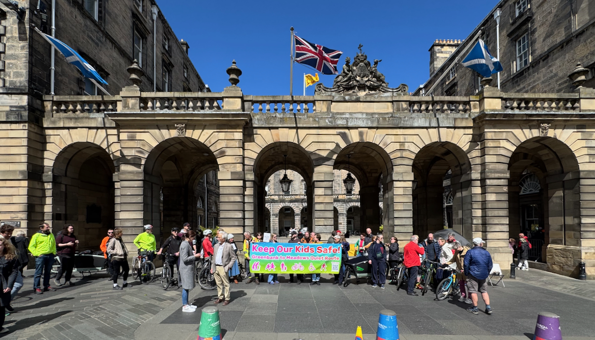 A mass of people gather in the sunshine outside of a civic building with a banner that reads 'keep our kids safe'