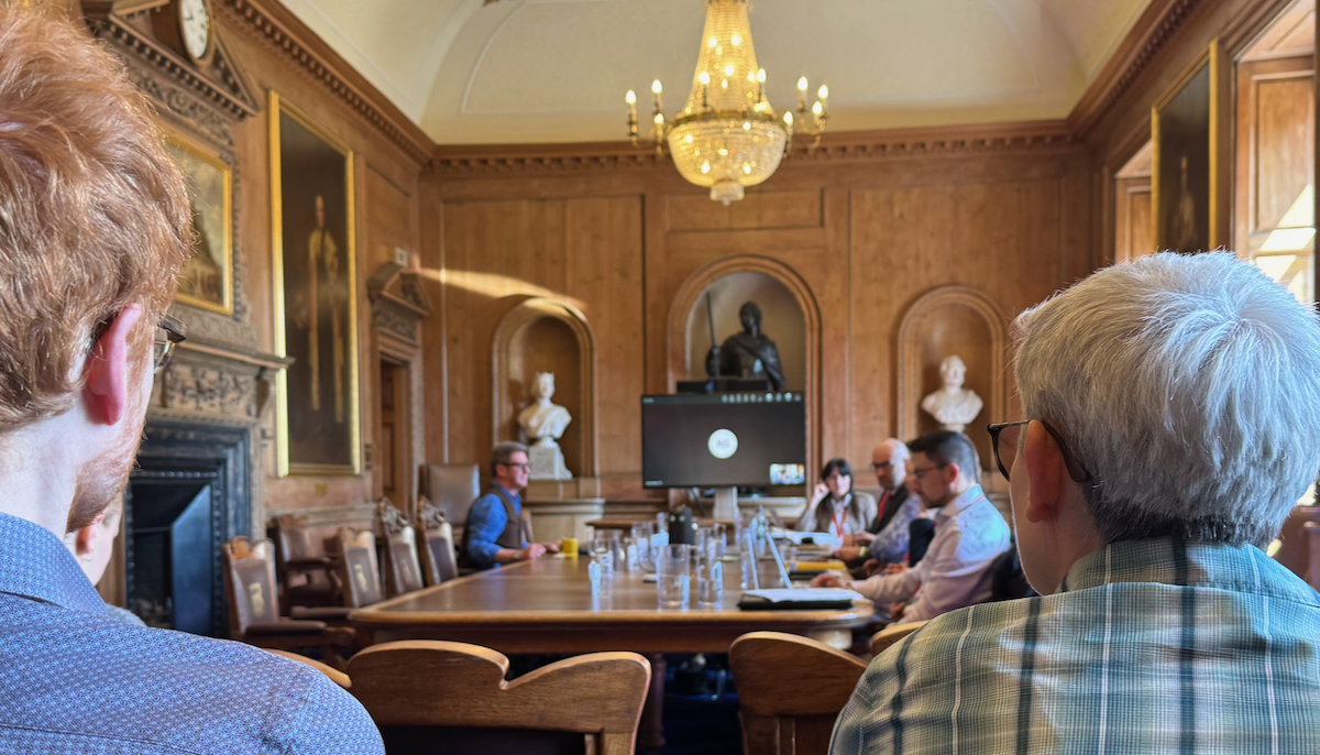 A grand meeting room flanked by oil paintings and wooden panelling hosts a chandelier or three and a large meeting table with a TV screen for members joining remotely