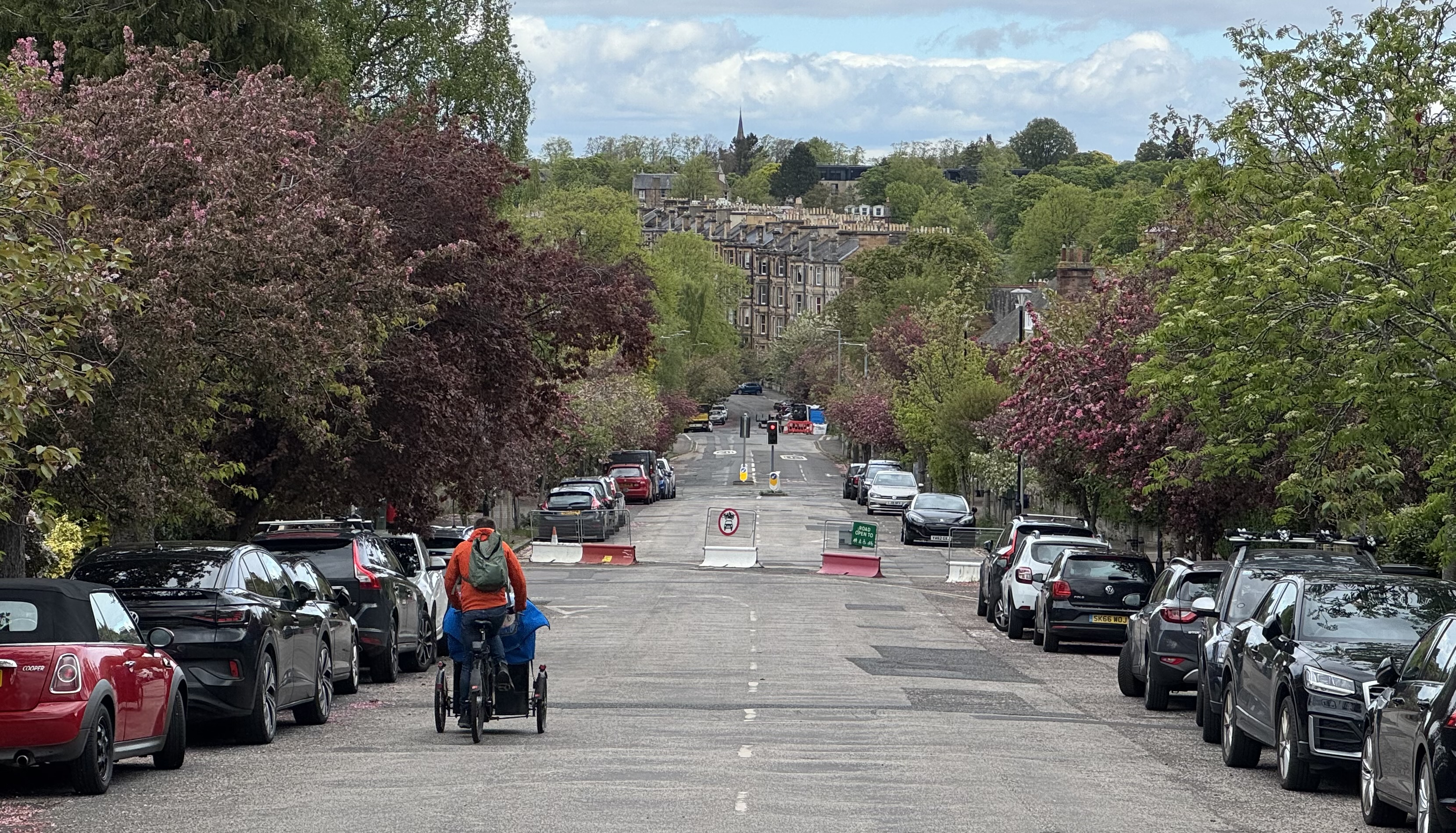 A parent rides a cargo trike with child onboard down a wide road towards a modal filter that is comprised of red and white metal fencing units spanning a junction
