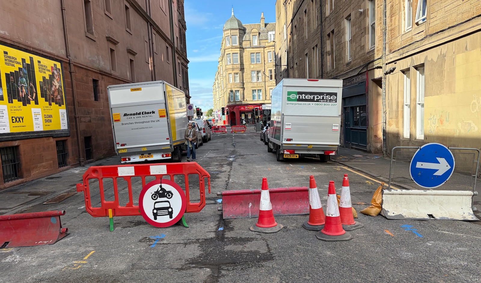 A photograph of an urban street blocked with a clutter of redirection signage and traffic cones, meanwhile beyond the barrier cars and vans have accessed and remain in place regardless